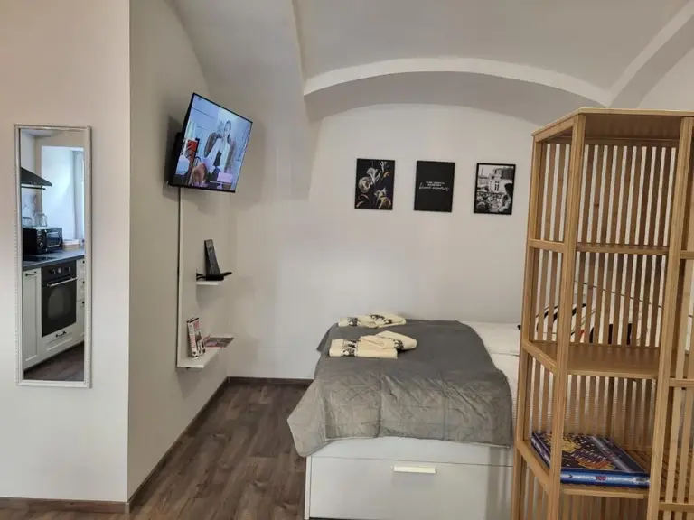 bedroom with gray bedspread, folded towels, wall-mounted TV, wooden shelving unit, and kitchen reflected in mirror